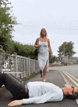 a woman in a dress is walking past a man laying on the sidewalk