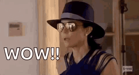 a woman wearing a hat and sunglasses is standing in front of a window and says wow .