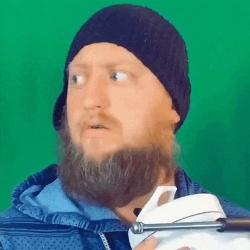 a man with a beard is wearing a hat and holding a white object in front of a green screen .