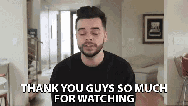 a man with a beard is standing in a living room and saying `` thank you guys so much for watching ''