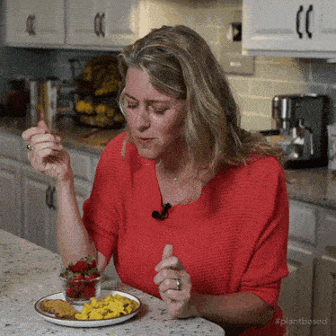 a woman in a red sweater sits at a counter eating a plate of food with the hashtag plantbased