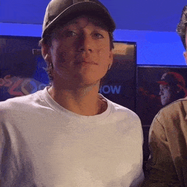 a man wearing a hat and a white shirt stands in front of a screen that says now