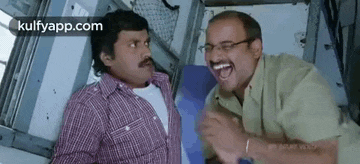two men are laughing on a train while sitting next to each other .