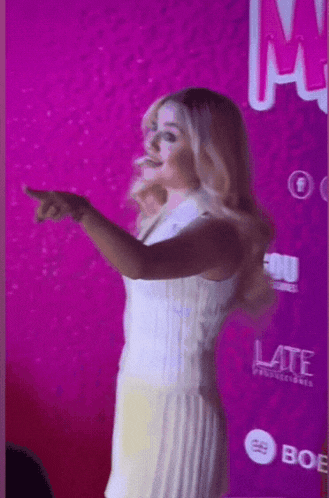 a woman in a white dress is pointing at something in front of a pink wall that says late