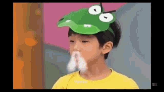 a young boy wearing a green hat with a frog on it is blowing his nose .