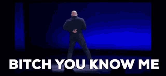 a man is standing on a stage with the words " bitch you know me " written on the screen behind him .