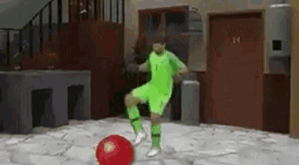 a man in a green jersey is kicking a red soccer ball on a sidewalk .