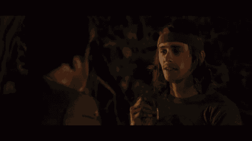 a man with long hair is smoking a cigarette while another man looks on