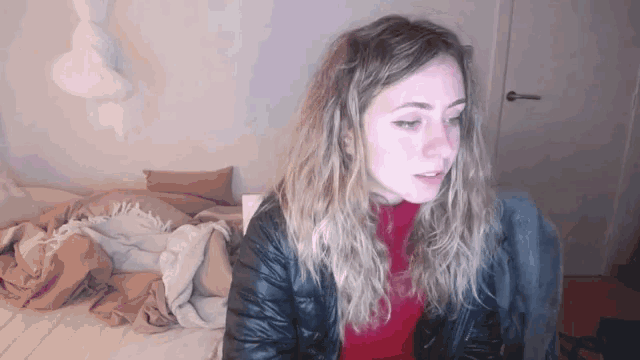 a woman in a black jacket sits on a bed with a blanket