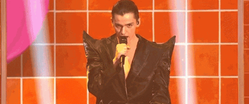 a man in a black jacket is singing into a microphone while standing in front of an orange tiled wall .