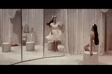 a woman is sitting on a stool in front of a mirror while another woman is dancing in a room .