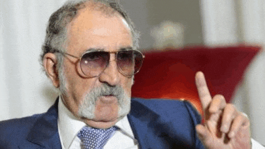 a man with a beard and sunglasses is sitting in a chair and pointing at something .