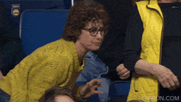 a woman wearing glasses and a yellow sweater is sitting in a ncaa stadium