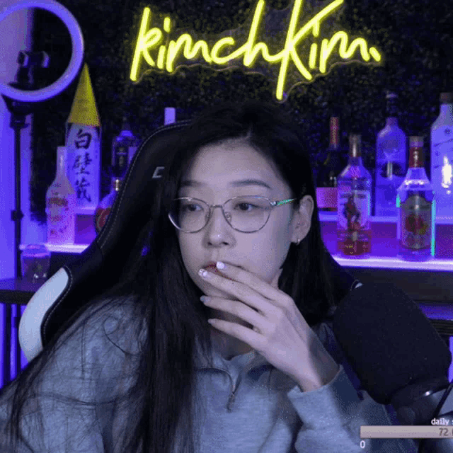 a woman wearing glasses is covering her mouth in front of a neon sign that says kimchikim
