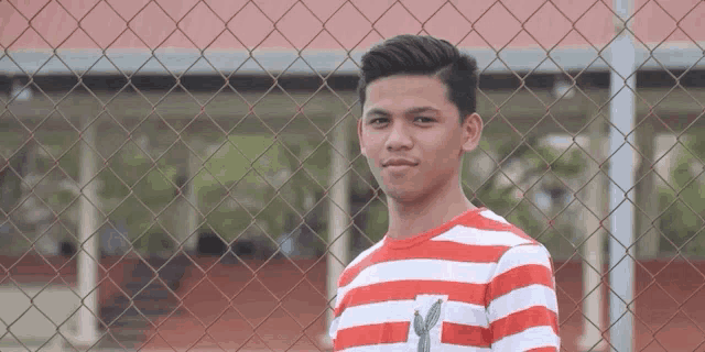 a young man wearing a red and white striped shirt is standing in front of a chain link fence