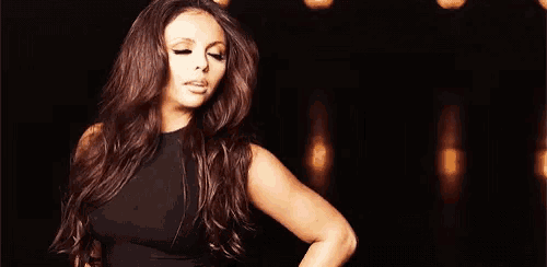 a woman with long brown hair is wearing a black dress and standing in front of a dark background .