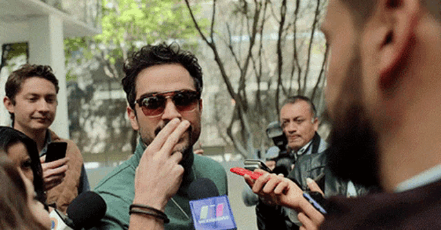 a man wearing sunglasses is covering his mouth with his hand while talking into a microphone that says ' noticias '