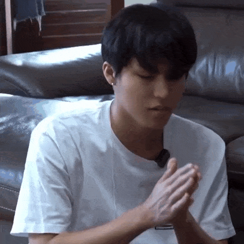 a young man in a white t-shirt is sitting on a couch with his hands together .