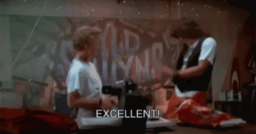 two men are sitting on the floor in front of a wall with graffiti on it and one of them is saying `` excellent '' .