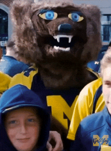 a boy wearing a blue hoodie with the letter m on it stands next to a mascot