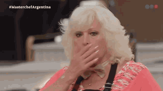 a woman with blonde hair is covering her mouth with her hand while watching a show called masterchef argentina