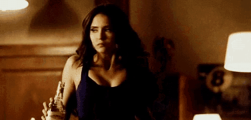 a woman in a blue tank top is holding a bottle of beer in her hand in a dark room .