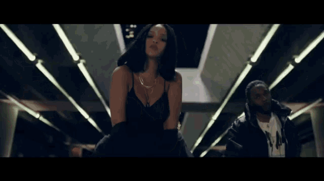 a woman in a black dress is dancing in a dark room with a man in a suit behind her .