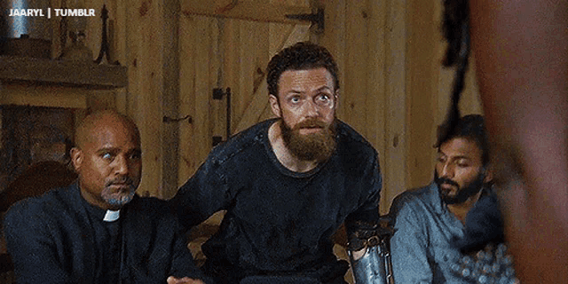 a man with a beard and a priest are sitting in a room with other men .