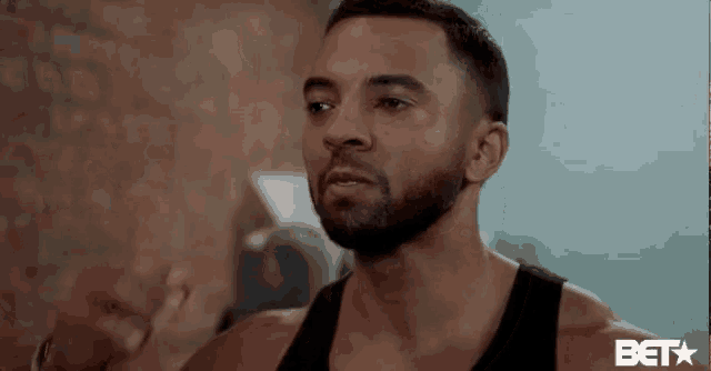 a man with a beard is wearing a black tank top and talking to someone .