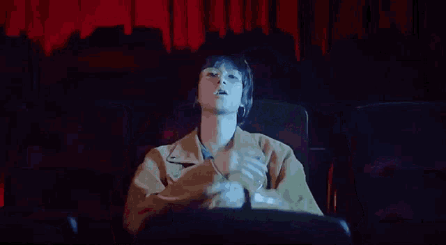 a man wearing glasses is sitting in a dark room with his hands up .