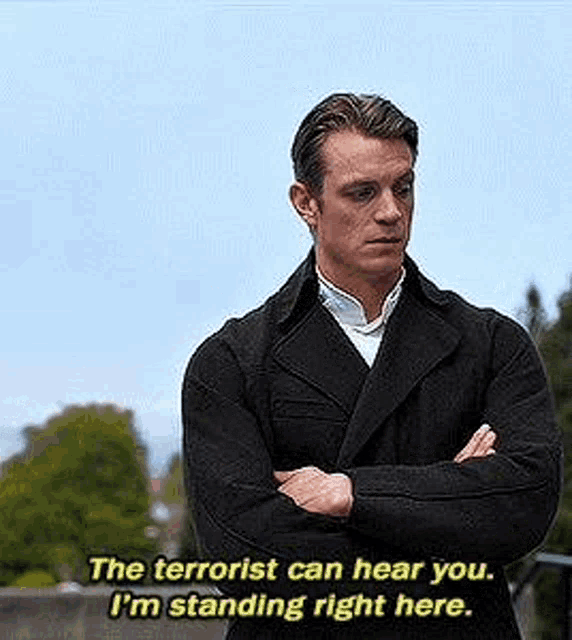 a man in a black coat is standing with his arms crossed and says `` the terrorist can hear you . ''