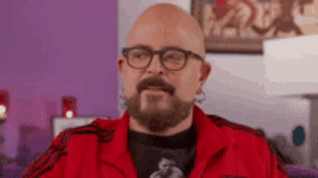 a bald man with a beard and glasses wearing a red jacket is sitting on a couch .