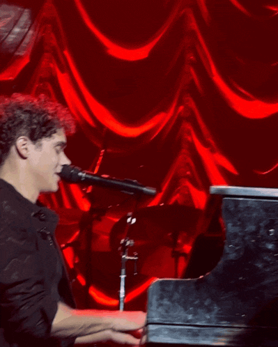 a man in a black shirt is playing a piano in front of a red curtain