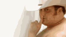 a shirtless man wearing a cowboy hat is holding his finger to his mouth .
