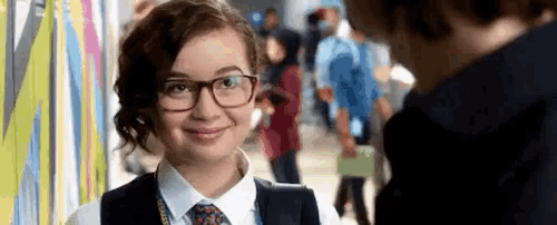 a young woman wearing glasses and a tie is smiling at a man in a hallway .