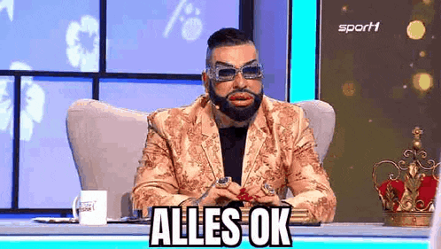 a man in a suit and sunglasses is sitting in a chair with a crown on his head and says alles ok .