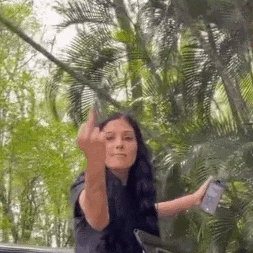 a woman is giving the middle finger while holding a cell phone in front of a forest .