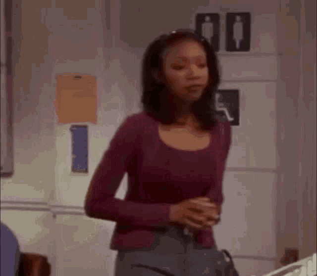 a woman in a purple shirt and gray pants is standing in front of a bathroom sign .