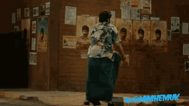 a man is walking in front of a wall with posters on it and the words iamhemuk on the bottom right