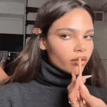 a woman wearing a black turtleneck is holding her finger to her lips .