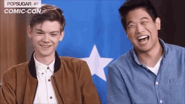 two young men are laughing in front of a blue and white flag with the words popsugar at comic con on it
