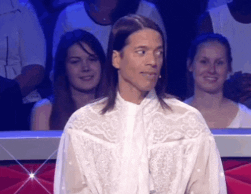 a man with long hair wearing a white shirt is sitting in a crowd .