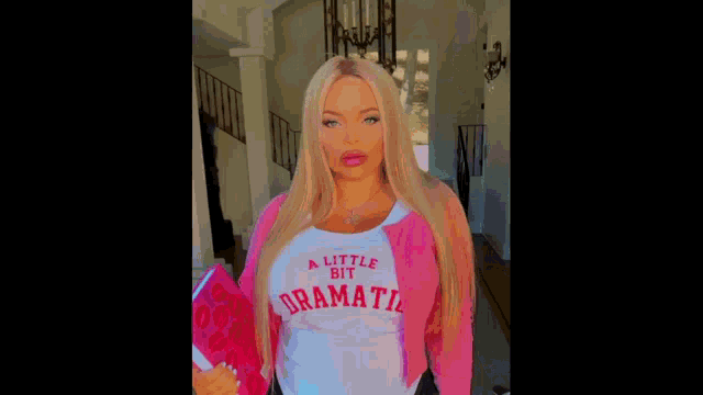 a woman with blonde hair is wearing a pink jacket and a white shirt that says `` a little bit dramatic '' .
