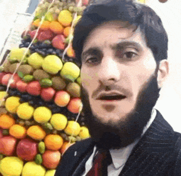 a man with a beard is standing in front of a stack of fruit .