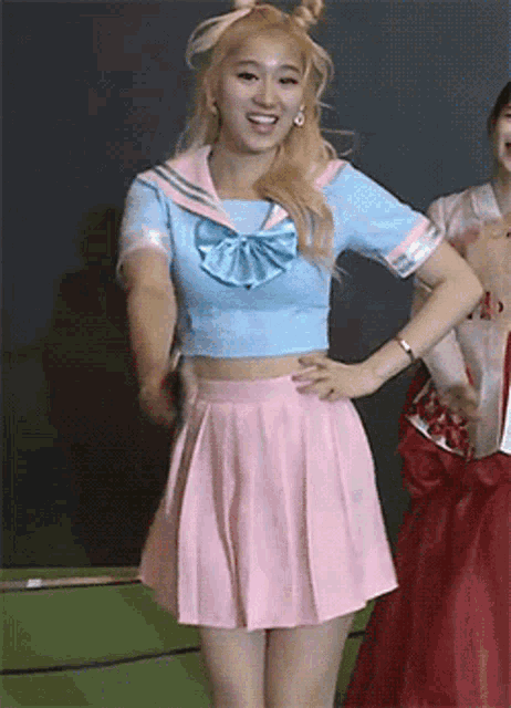 a woman wearing a blue top and a pink pleated skirt stands with her hands on her hips