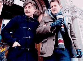 two men are standing next to each other in front of a building . one of the men is holding a cup of coffee .