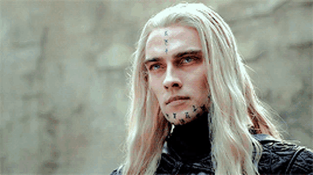 a man with long blonde hair has a tattoo on his face .
