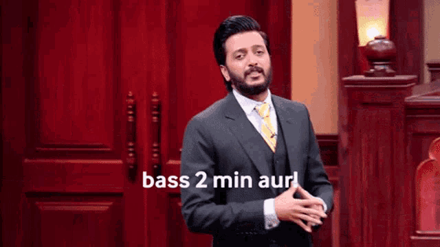 a man in a suit and tie is standing in front of a wooden door and says bass 2 min aur