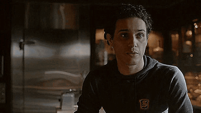 a young man wearing a black sweater with a shield on it is standing in a dark room .