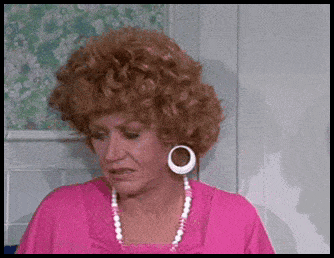 a woman with a very large wig and earrings is wearing a pink shirt and a pearl necklace .